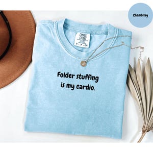 May include: A pale blue t-shirt with the slogan "Folder stuffing is my cardio" in black lettering. The shirt has a crew neck and short sleeves. A silver necklace with a pendant is draped across the shirt. The shirt is labeled "Chambray".