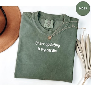 Pode incluir: Uma camiseta Comfort Colors verde musgo com o texto "Chart updating is my cardio" em branco. Um colar de ouro repousa sobre a camiseta. A camiseta está dobrada sobre uma superfície branca.