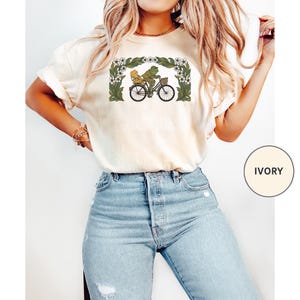 Puede incluir: Camiseta de color marfil con una ilustraci&oacute;n caprichosa de una rana y un acompa&ntilde;ante en bicicleta, enmarcada por un borde floral. El dise&ntilde;o es en tonos verdes, amarillos y negros. La camiseta es una prenda informal.