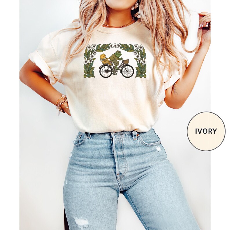 Puede incluir: Camiseta de color marfil con una ilustraci&oacute;n caprichosa de una rana y un acompa&ntilde;ante en bicicleta, enmarcada por un borde floral. El dise&ntilde;o es en tonos verdes, amarillos y negros. La camiseta es una prenda informal.