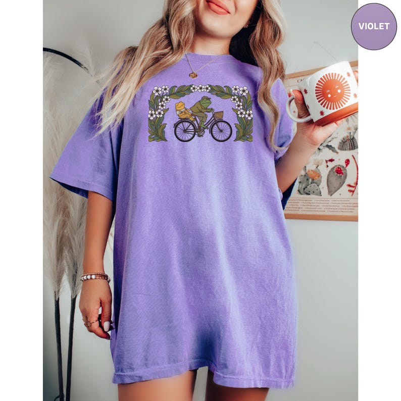 Puede incluir: Una camiseta de color lila con una ilustraci&oacute;n caprichosa de dos ranas montando en bicicleta, enmarcada por flores y follaje. El dise&ntilde;o est&aacute; centrado en la camiseta, que tiene mangas cortas y un corte holgado. La camiseta est&aacute; siendo sostenida por una persona.