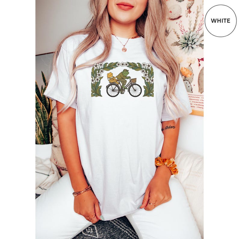 Puede incluir: Camiseta blanca con una ilustraci&oacute;n caprichosa de una rana montando en bicicleta con un pasajero. El dise&ntilde;o est&aacute; enmarcado por un borde floral en verde y blanco. La bicicleta tiene una cesta. La camiseta es una prenda casual.