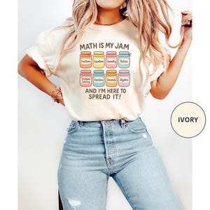 Puede incluir: Camiseta de color marfil con el texto "MATH IS MY JAM" sobre botes coloridos con términos matemáticos como Fracciones y Álgebra. La frase "AND I'M HERE TO SPREAD IT!" está debajo. La camiseta se combina con vaqueros azules.