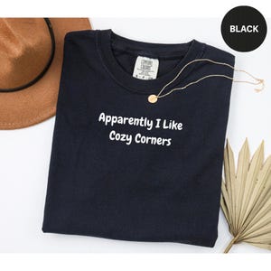 May include: Black t-shirt with the text "Apparently I Like Cozy Corners" in white. A gold necklace and a brown hat are next to the shirt. A black circle with the word "BLACK" is in the upper right corner.
