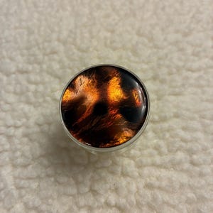 May include: A round bottle stopper featuring a tortoiseshell design in brown, black, and amber hues. It has a silver-coloured metal rim and a glossy finish. The pattern is reminiscent of flames or an abstract, fiery design.
