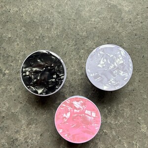 May include: Three round phone grips in black, white, and pink. Each grip has a pearlescent, textured surface and a white rim. The grips are arranged on a grey, speckled surface.