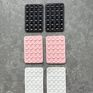 May include: Six rectangular silicone pads in black, pink, and white. Each pad features a grid of small, rounded bumps. The pads are arranged in pairs, with two of each colour visible. These pads are likely designed for grip or non-slip purposes.