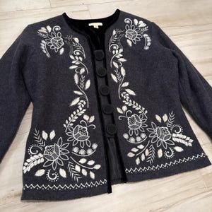 May include: A dark gray jacket with white floral embroidery and black velvet trim. The jacket features a round collar, long sleeves, and a row of black buttons down the front. The floral design includes leaves and flowers.
