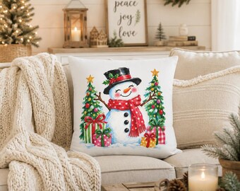 May include: A white square throw pillow featuring a central illustration of a smiling snowman wearing a black top hat and a red polka-dot scarf, flanked by two decorated Christmas trees and colourful gift boxes on a snowy ground base.