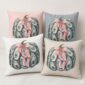 May include: Four square throw pillows featuring a central pumpkin illustration composed of blue and white oyster shells, tied with a soft pink ribbon. The pillows have solid backgrounds in light pink, light blue, white, and beige, respectively.