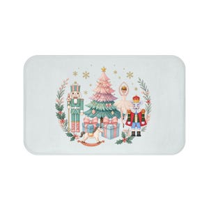 May include: A rectangular bath mat featuring a central illustration of a pink and green Christmas tree, a nutcracker, a ballerina, a mouse king, a rocking horse, and gift boxes, all surrounded by a wreath of holly leaves and berries.