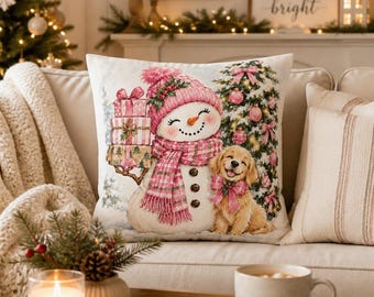 May include: A square throw pillow featuring a printed illustration of a smiling snowman wearing a pink knit hat and scarf, holding a stack of three wrapped gifts, next to a golden retriever puppy and a decorated Christmas tree.