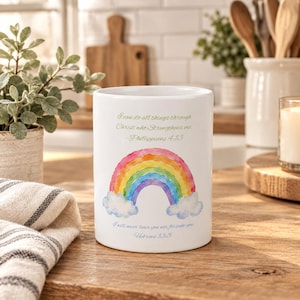 May include: A white ceramic mug featuring a watercolor rainbow graphic between two clouds. The text reads: I can do all things through Christ who strengthens me. Philippians 4:13 and I will never leave you nor forsake you. Hebrews 13:5.