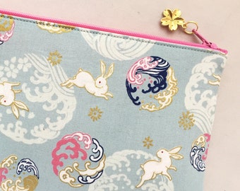 May include: A light blue zippered pouch with a pink zipper and a gold charm. The pouch has a repeating pattern of white rabbits, blue and gold waves, and pink and gold circles.