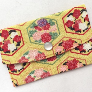May include: A yellow fabric wallet with a floral pattern and a silver button closure. The wallet has a hexagonal design with red, white, and green flowers.