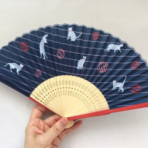 May include: A blue and white hand fan with a red trim. The fan features a repeating pattern of white cats and red balls on a blue striped background. The fan is made of paper and has a wooden handle.