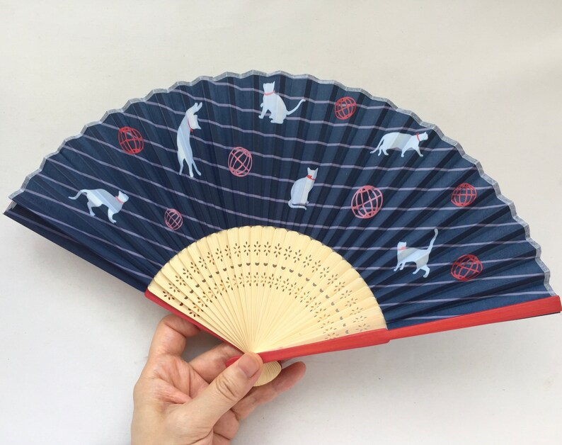 May include: A blue and white hand fan with a red trim. The fan features a repeating pattern of white cats and red balls on a blue striped background. The fan is made of paper and has a wooden handle.