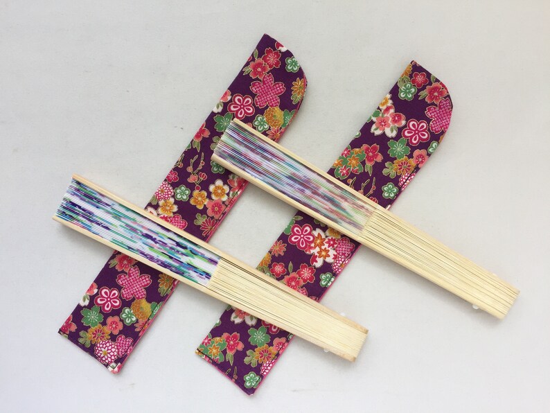 May include: Three folding fans with purple fabric covers featuring a floral pattern. The fans are made of wood and have a white and blue patterned paper insert.