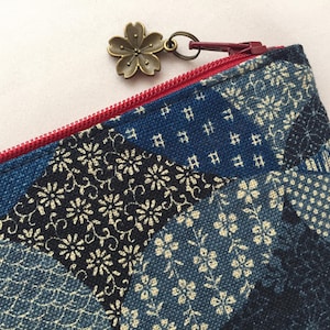 May include: Close-up of a blue and white patterned pouch with a red zipper. The pouch features a floral design and a bronze-colored flower charm attached to the zipper pull. The fabric has a patchwork design.