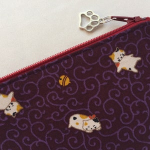 May include: A purple zippered pouch with a paw print zipper pull. The pouch is decorated with a repeating pattern of white cats and yellow bells on a purple background.