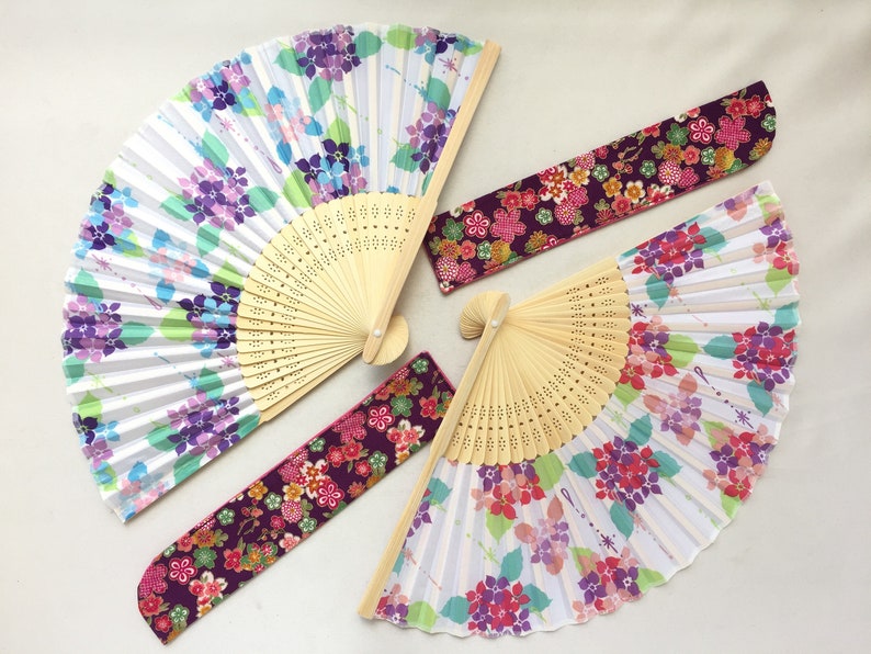 May include: Four folding fans with floral designs. Two fans have a white background with purple and green flowers. One fan has a purple background with pink and red flowers. The last fan has a purple background with pink, red, and white flowers.