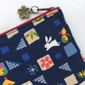May include: A blue zippered pouch with a white rabbit, flowers, and other Japanese-inspired patterns. The pouch has a red zipper and a small bronze charm attached to the zipper.