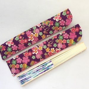 May include: Two purple fabric fan covers with pink and yellow floral patterns. The covers are folded and placed on top of a wooden fan with a white and blue patterned paper.