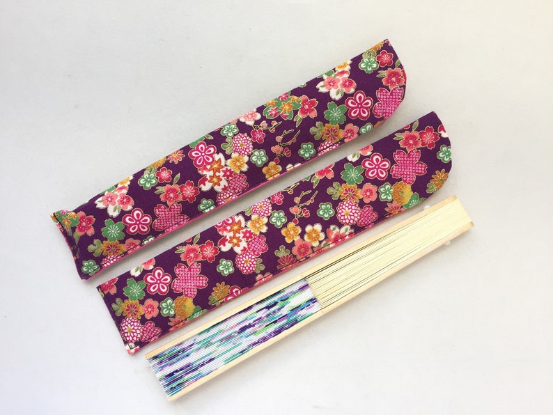 May include: Two purple fabric fan covers with pink and yellow floral patterns. The covers are folded and placed on top of a wooden fan with a white and blue patterned paper.