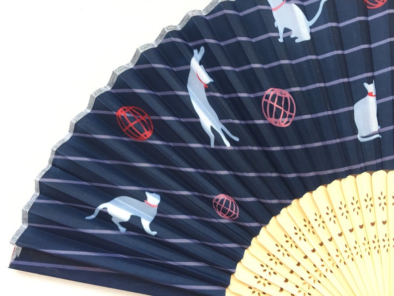 May include: A blue and white folding fan with a striped pattern and a white cat design. The fan has a wooden handle with a decorative pattern.