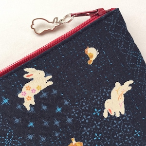 May include: A dark navy blue fabric pouch featuring a repeating pattern of cream-colored rabbits and small floral accents. The pouch has a bright red zipper closure with a silver-toned metal rabbit-shaped charm attached to the zipper pull.