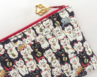May include: A black and white zippered pouch with a red zipper pull. The pouch is covered in a pattern of white cats with raised paws, black cats, and red bells. The cats are all facing forward and have a happy expression.