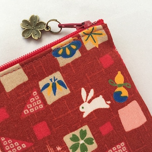 May include: A red zippered pouch with a checkered pattern and various colorful designs, including a rabbit, a flower, and a gourd. The pouch has a gold metal charm attached to the zipper.