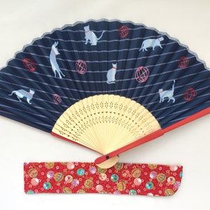 May include: A blue and white folding fan with a red handle and a red and white fabric case. The fan has a striped design with cats and red balls printed on it.