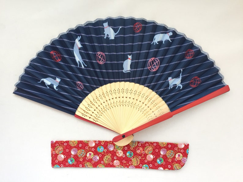 May include: A blue and white folding fan with a red handle and a red and white fabric case. The fan has a striped design with cats and red balls printed on it.