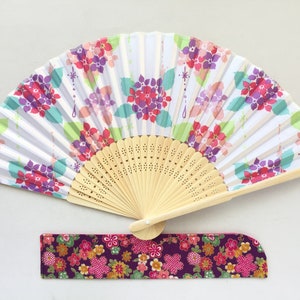 May include: A white folding fan with a floral design in shades of pink, purple, and green. The fan has a wooden handle and a matching fabric pouch.