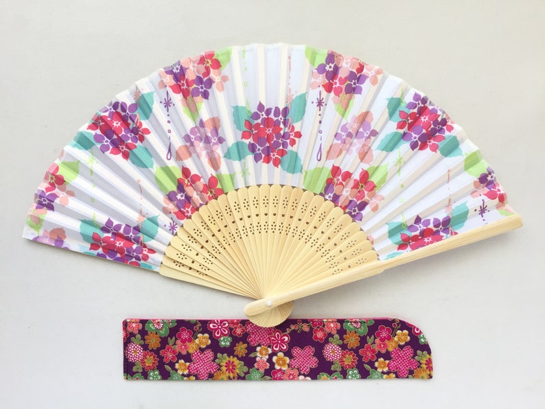 May include: A white folding fan with a floral design in shades of pink, purple, and green. The fan has a wooden handle and a matching fabric pouch.