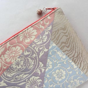 May include: A colorful, patterned pouch with a red zipper. The pouch features sections of pink, lavender, and blue fabric with floral designs, and a section of beige fabric with a textured pattern. A small, beaded charm hangs from the zipper.