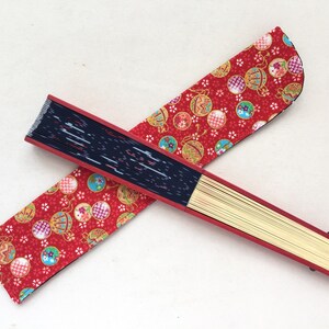 May include: A red and black folding fan with a patterned fabric cover. The fabric features a repeating design of red, gold, and blue ornaments on a red background.