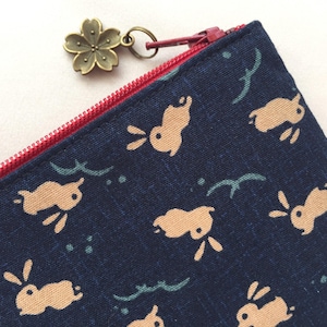 May include: A dark blue fabric pouch featuring a repeating pattern of light tan rabbits and small teal wave motifs. The pouch has a bright red zipper closure and a metallic bronze-toned zipper pull shaped like a five-petaled cherry blossom flower.