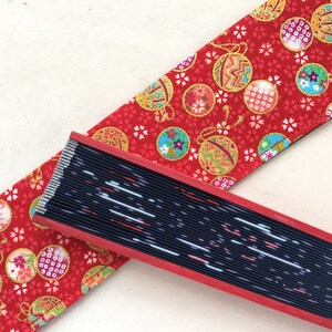 May include: A red fabric with a pattern of gold and blue ornaments on a red background. A black and red folding fan with a white and blue design is lying on top of the fabric.