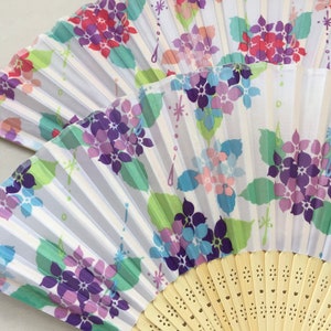 May include: A white folding fan with a floral design. The fan features purple, pink, and blue flowers with green leaves on a white background. The fan is made of paper and has a wooden handle.