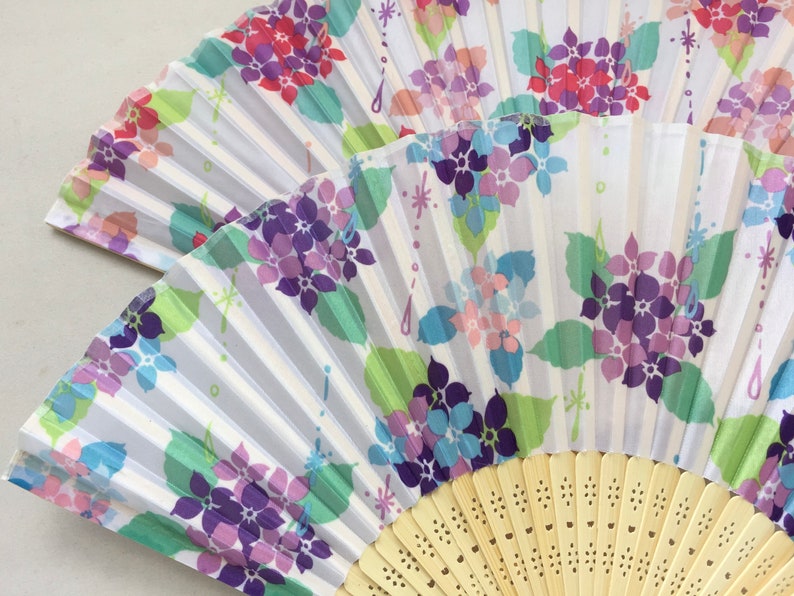 May include: A white folding fan with a floral design. The fan features purple, pink, and blue flowers with green leaves on a white background. The fan is made of paper and has a wooden handle.