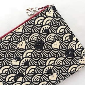 May include: A black and cream-colored zippered pouch with a repeating pattern of stylized cats and wave-like shapes. The zipper is red, and a silver flower charm hangs from the zipper pull.