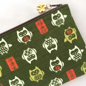 May include: Green fabric pencil case with a repeating pattern of white and yellow owls. The owls have various designs and are interspersed with red rectangular stamps. A gold-colored flower charm hangs from the zipper.