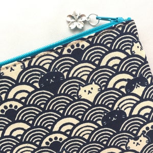 May include: A black and white zippered pouch with a blue zipper pull. The pouch has a repeating pattern of black cats and paw prints on a white background. The pattern is set against a background of black and white waves.