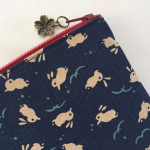 May include: A dark blue zippered pouch with a repeating pattern of beige bunnies and green leaves. The zipper pull is a small bronze flower charm.