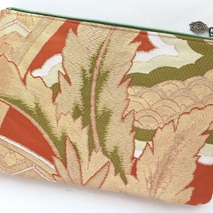 May include: A zippered pouch with a green zipper and a gold charm. The pouch is made of a fabric with a woven pattern of orange, green, and gold leaves.