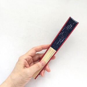May include: A red and blue hand fan with a wooden frame. The fan is closed and being held by a hand.