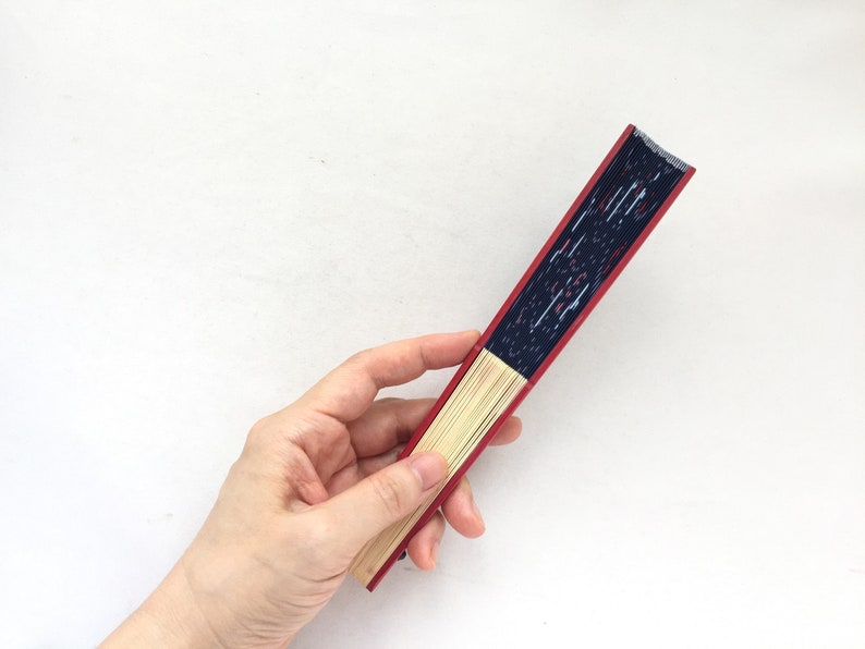May include: A red and blue hand fan with a wooden frame. The fan is closed and being held by a hand.