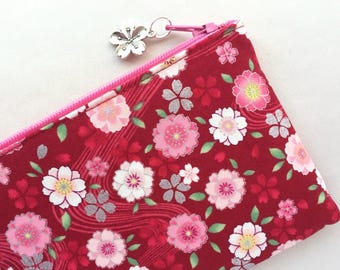 May include: A rectangular fabric pouch in a deep red color featuring a repeating pattern of pink and white cherry blossoms with small green leaves. The pouch has a bright pink zipper closure with a silver-toned metal cherry blossom charm attached.
